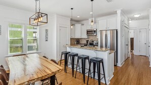 A modern kitchen with white cabinetry, a wooden dining table, bar stools, pendant lighting, and a stainless steel refrigerator. - Gorgeous Sound-side Family Friendly Retreat l Golf Cart Included l Convenient to National Seashore (Navarre)