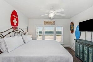 Ein Schlafzimmer mit Bett, Deckenventilator, Fernseher und Meerblick durch große Fenster.