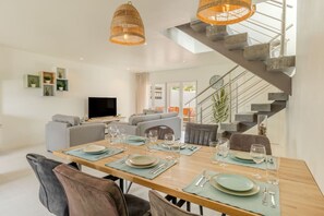 A modern dining area with a wooden table set for four, surrounded by comfortable seating. A staircase with a metal railing leads to an upper level.