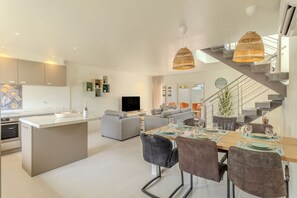 Une cuisine moderne dotée d’un îlot central, un coin repas avec une table et des chaises en bois, un salon avec des canapés et un escalier avec une rampe en verre.