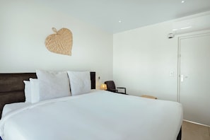 A hotel room with a large bed, a dark headboard, a wall-mounted heart-shaped decoration, a bedside table with a lamp, and a door.