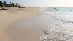 Une vaste plage de sable fin, avec des vagues douces, des palmiers et des gens qui profitent de l’eau.