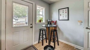 A kitchen with a white door, two windows, a small table with stools, and a framed menu on the wall.