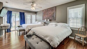 A bedroom with two beds, a ceiling fan, a large window with blinds, and a painting on the wall.