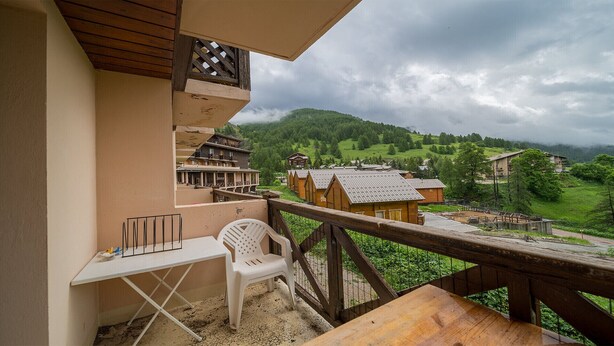 Un balcone con un tavolo di legno e una sedia di plastica bianca, affaccia su un villaggio con case tradizionali e un paesaggio montuoso.
