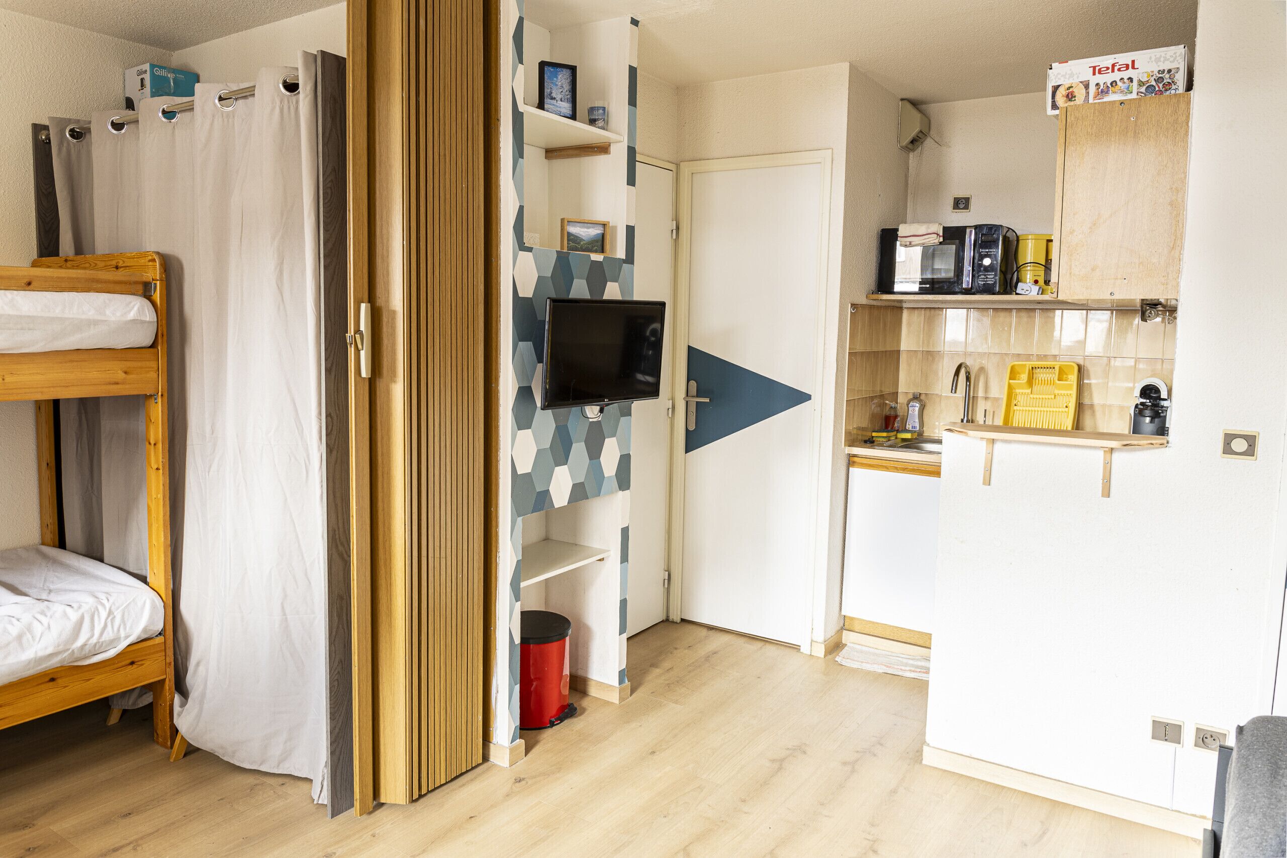 Une chambre avec un lit superposé, un téléviseur fixé au mur, un coin cuisine équipé d’un four à micro-ondes et d’un grille-pain, et un meuble portant le logo Tefal.