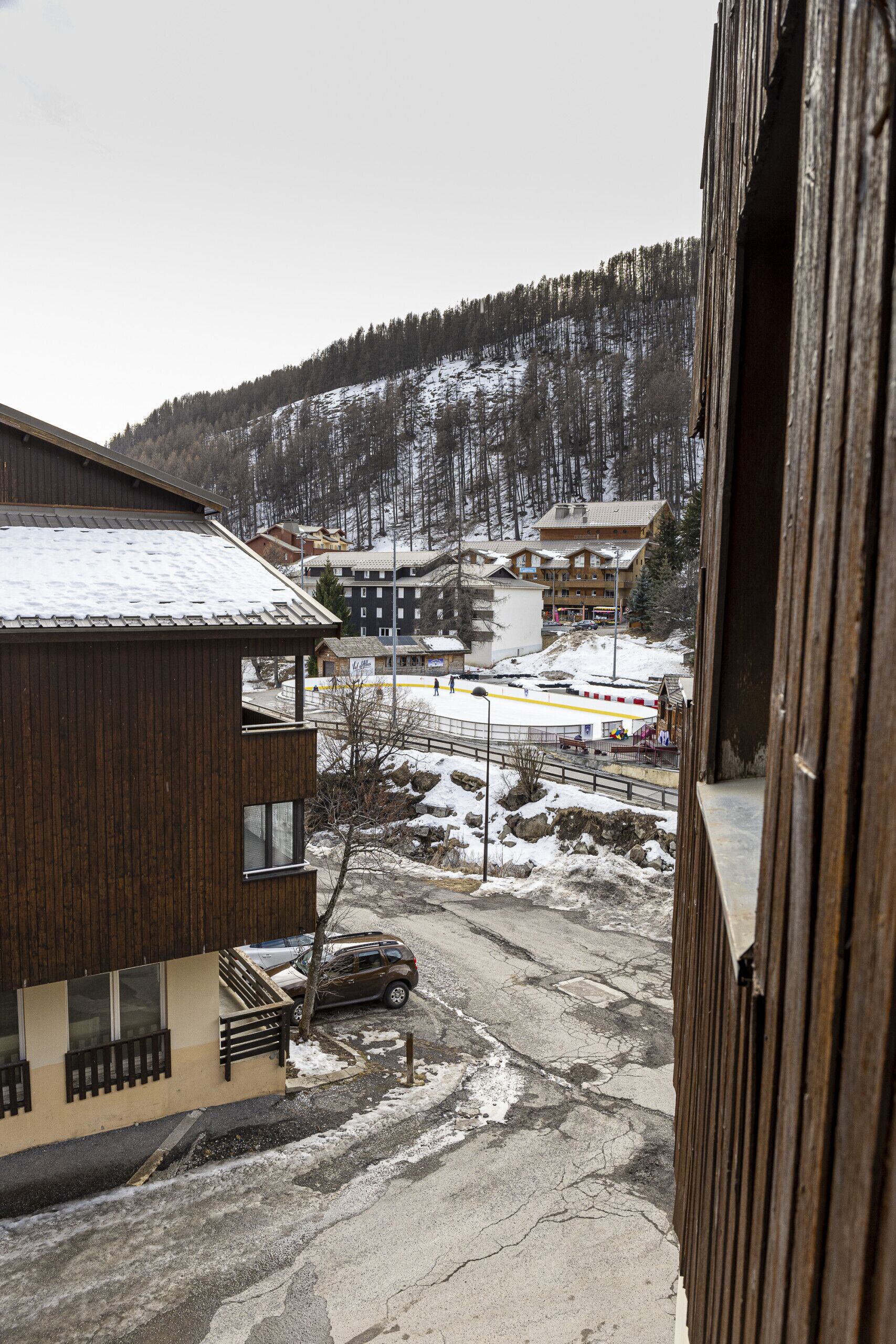 A wooden building with a balcony overlooking a snowy landscape, a parking area, and a distant view of a town with buildings and a forested hill.