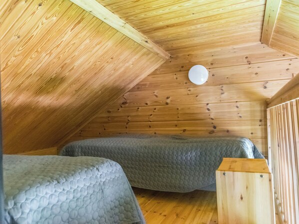 A small, cozy attic bedroom with wooden walls and a sloped ceiling. There are two beds with grey bedding and a small wooden nightstand.