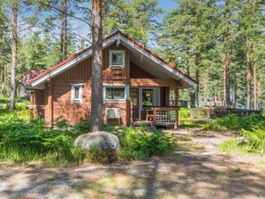 A wooden cabin with a porch, surrounded by trees and a gravel path.