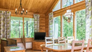 L’intérieur chaleureux d’un chalet comprend une table à manger, des chaises, un téléviseur et de grandes fenêtres donnant sur la forêt.