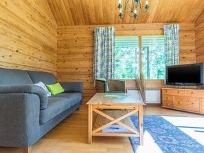A cozy cabin interior with a wooden ceiling, a gray sofa with a green pillow, a wooden table with a book and a green cloth, a TV on a wooden cabinet, and a window with curtains. - Vacation home Paju 7 by Interhome (Loviisa)
