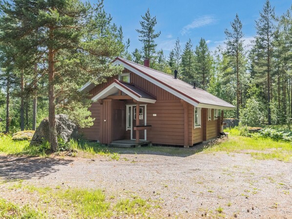 Une petite cabane dotée d’un porche à l’avant, entourée d’arbres et bordée d’une allée en gravier.