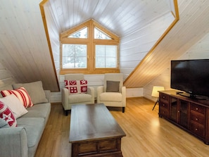 Acogedor dormitorio en ático con techo de madera, un ventanal grande, televisor y sofá con cojines decorativos.