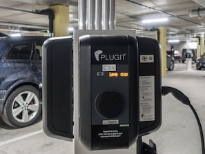 A parking station with a charging plug, a car parked nearby, and a QR code.