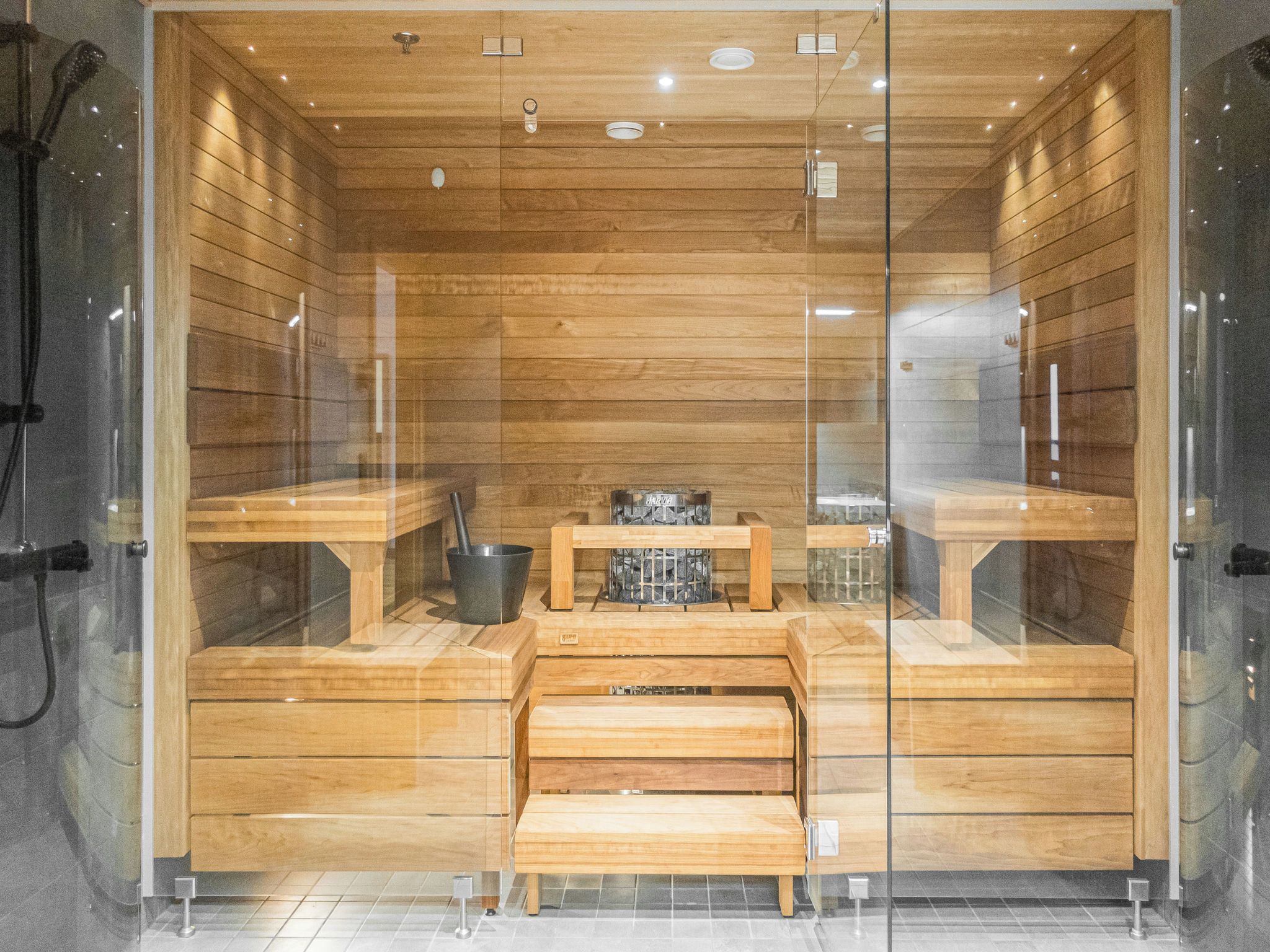 A sauna with wooden benches and a central table, glass walls, and a tiled floor.