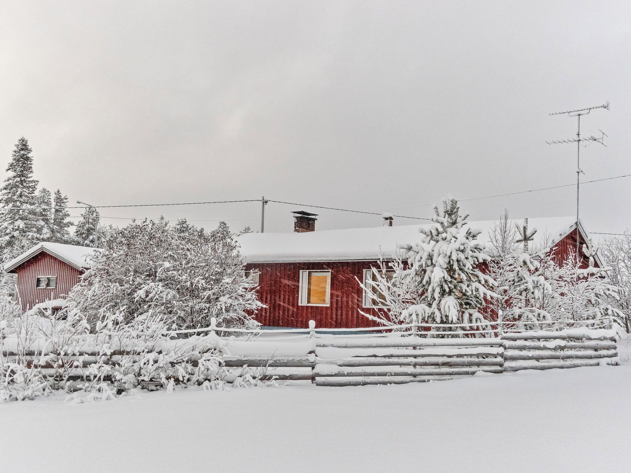 Ett rött trähus med ett väl synligt fönster och en skorsten, omgivet av snötäckta träd och ett staket.
