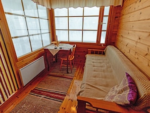 Un accogliente interno di una baita con pavimento in legno, un divano, un tavolo con una sedia e ampie finestre.