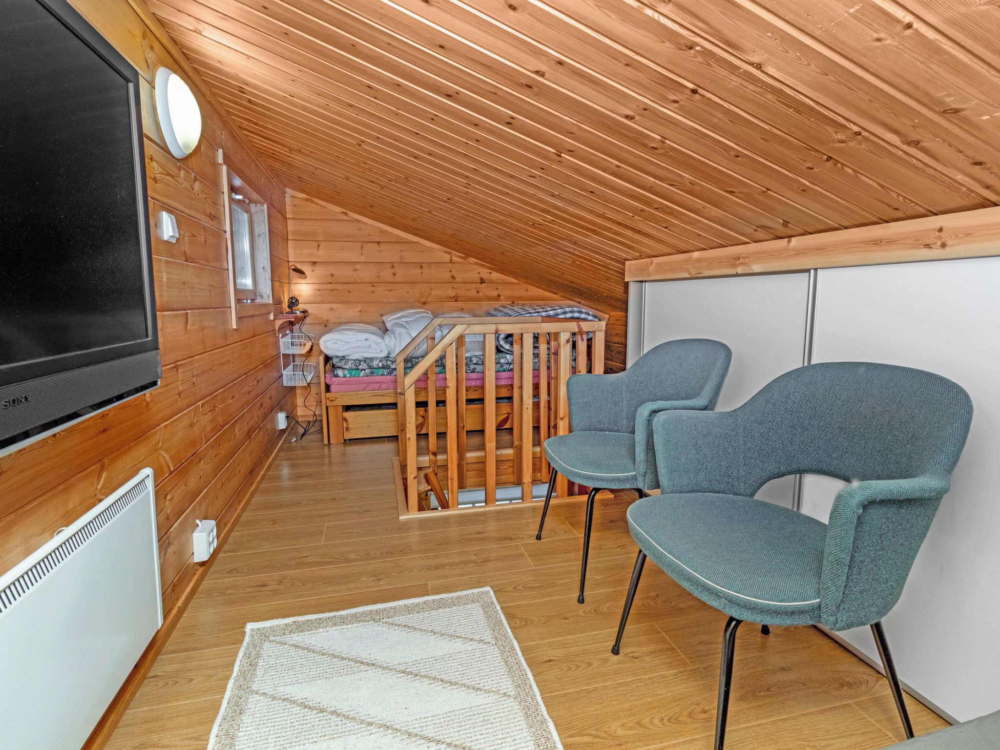 Acogedor dormitorio en ático con paredes de madera y techo inclinado. Se observa una cama con cabecera de madera, un televisor y dos sillas.