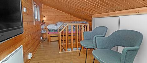 Acogedor dormitorio en ático con paredes de madera y techo inclinado. Se observa una cama con cabecera de madera, un televisor y dos sillas.
