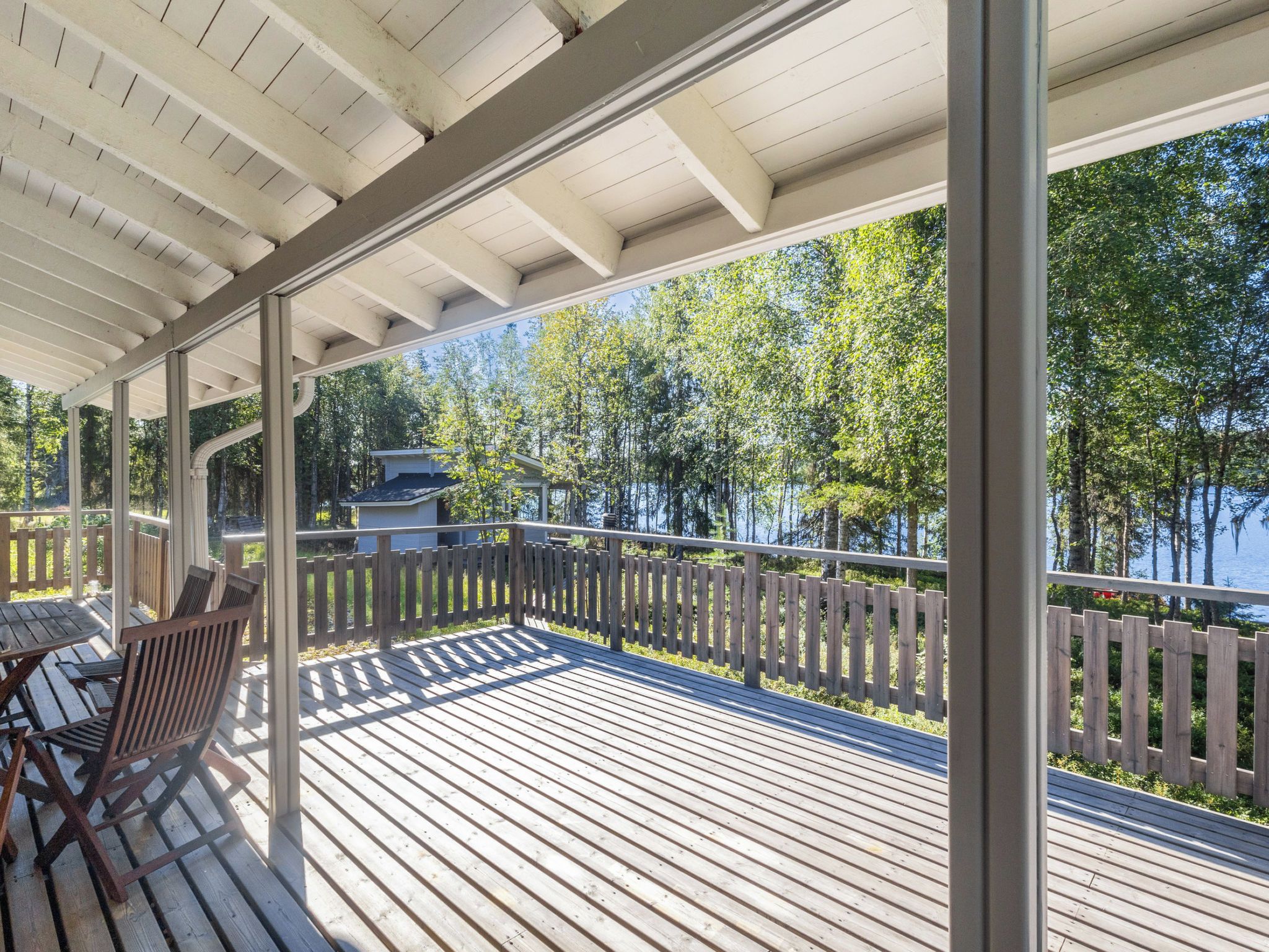 A covered deck with a view of a lake and trees, wooden furniture, and a wooden fence.