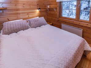 Une chambre dans une cabane en bois, avec un lit, des oreillers, une table de chevet, une étagère, une fenêtre donnant sur des arbres couverts de neige et un lampadaire fixé au mur.