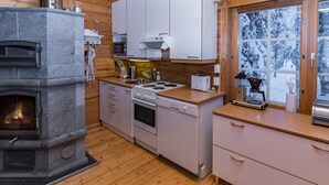 A modern kitchen with a stone fireplace, wooden cabinets, and a window with a snowy view. - Pohjaslahdentie 17 b by Interhome (Kuusamo)