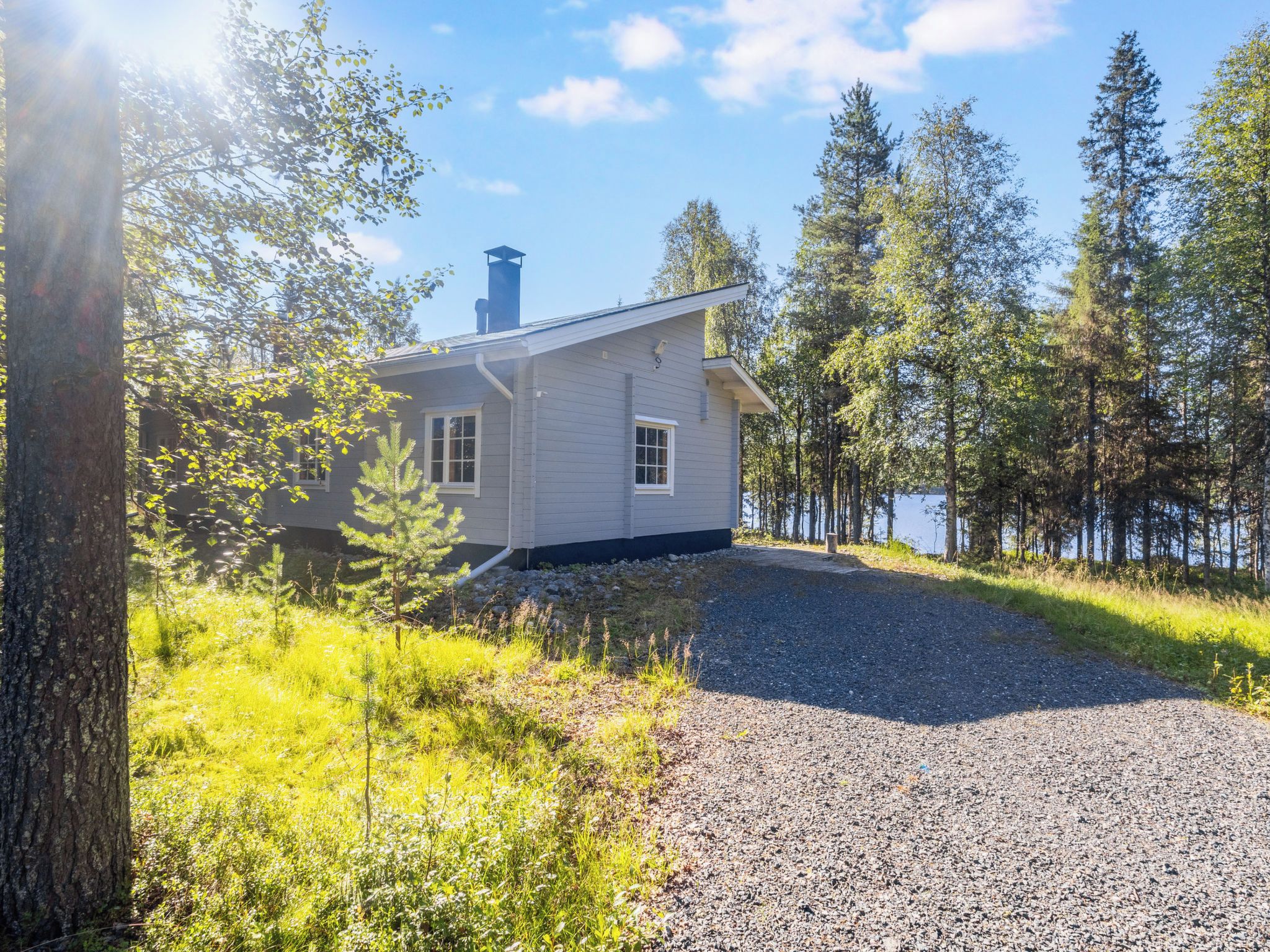 A house with a gravel driveway, surrounded by trees and a body of water.