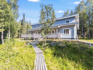 A two-story house with a deck and a covered outdoor area, surrounded by trees and a grassy landscape. - Pohjaslahdentie 17 b by Interhome (Kuusamo)