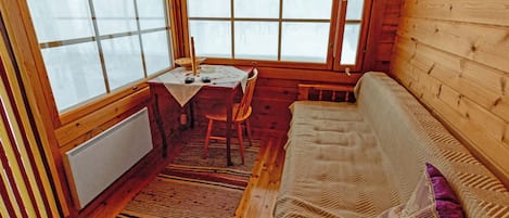 A cozy cabin interior with a wooden floor, a sofa, a table with a chair, and large windows.
