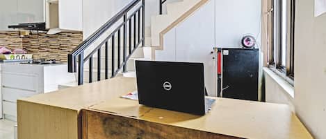 A room with a wooden desk, a laptop, a chair, a staircase, and a kitchen area.