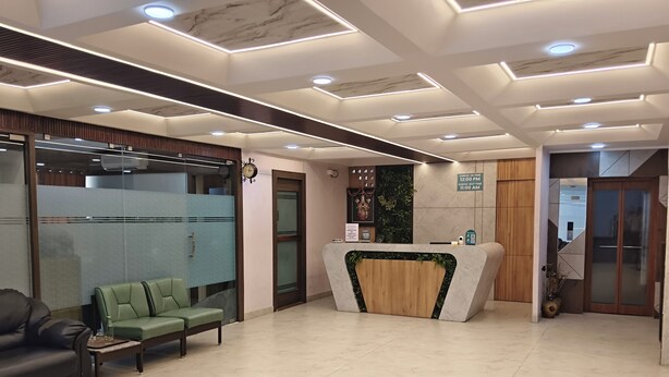 A reception area with a wooden reception desk, a green sofa, a black leather chair, and a tiled floor. - Navaratna Delight (Coimbatore)