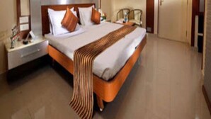 A hotel room with a large bed, a nightstand, a bedside table, a chair, and a door. - Navaratna Delight (Coimbatore)