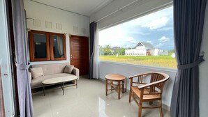 Una habitación con sofá, una mesita y dos sillas, un ventanal con cortinas y vistas a una casa y campos.