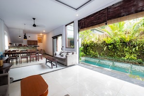 A modern living area with a dining set, a sofa, and a poolside view with lush greenery.