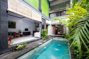 A modern indoor pool with a clear view of the surrounding area, including a seating area with a sofa and a dining table, and a balcony with plants. - Villa Kubu Arya by Supala (Ubud)