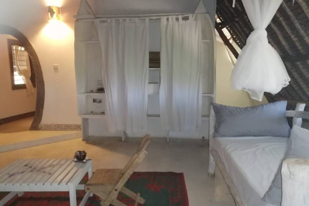 Ein Schlafzimmer mit einem Bett, einem Holzstuhl, einem kleinen Tisch, einem Nachttisch und einem Fenster mit Vorhängen.