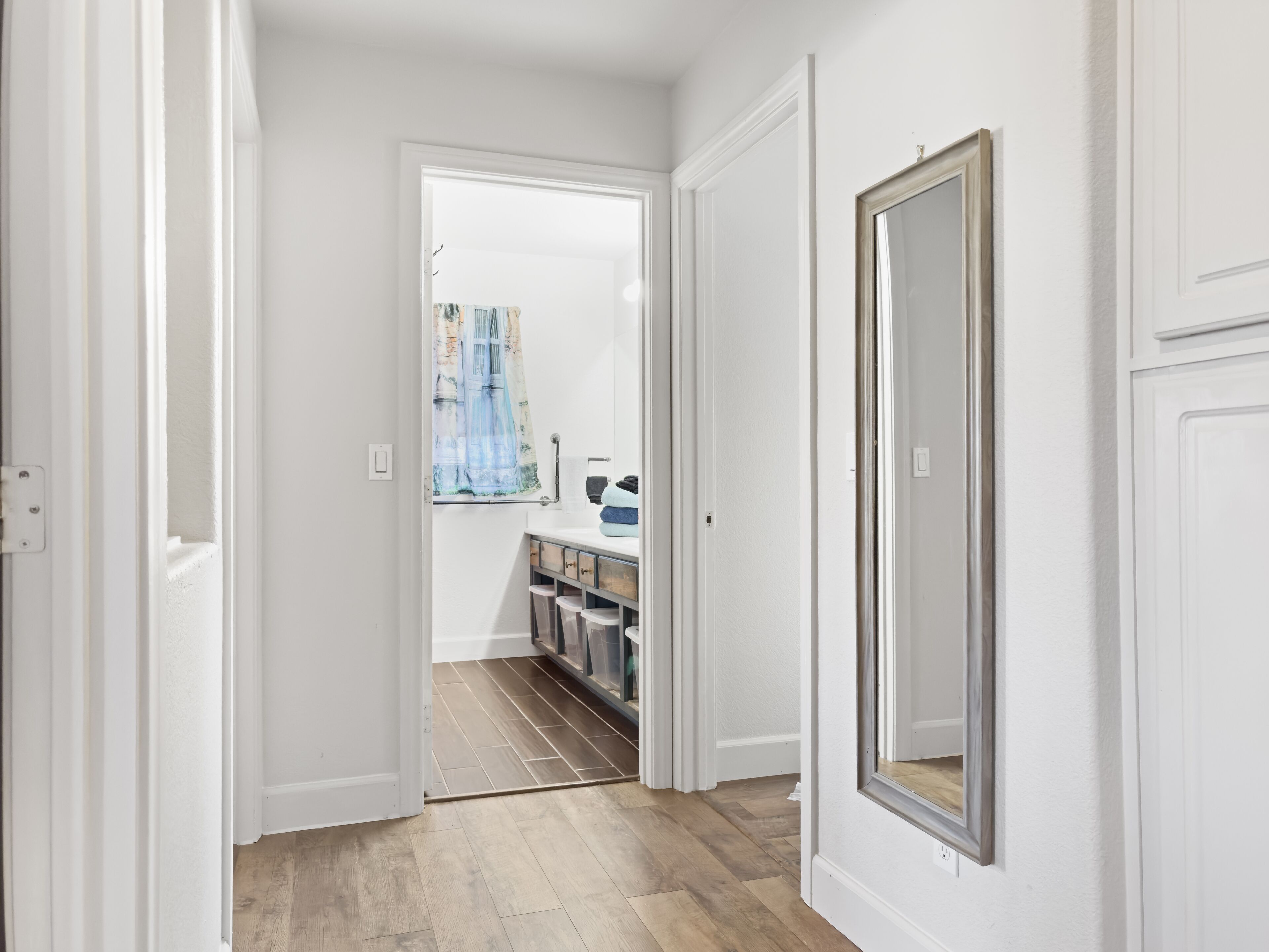 A hallway with white doors, a wooden floor, and a view into a room with a window and a shelf.
