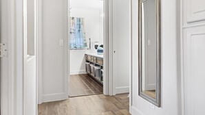 Un pasillo con puertas blancas, suelo de madera y vista a una habitación con ventana y estantería.