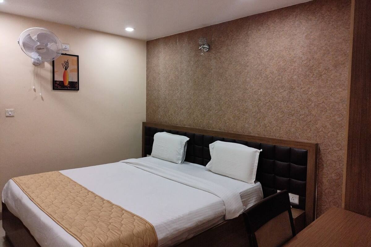 A hotel room with a bed, a chair, a table, a wall clock, and a ceiling fan.