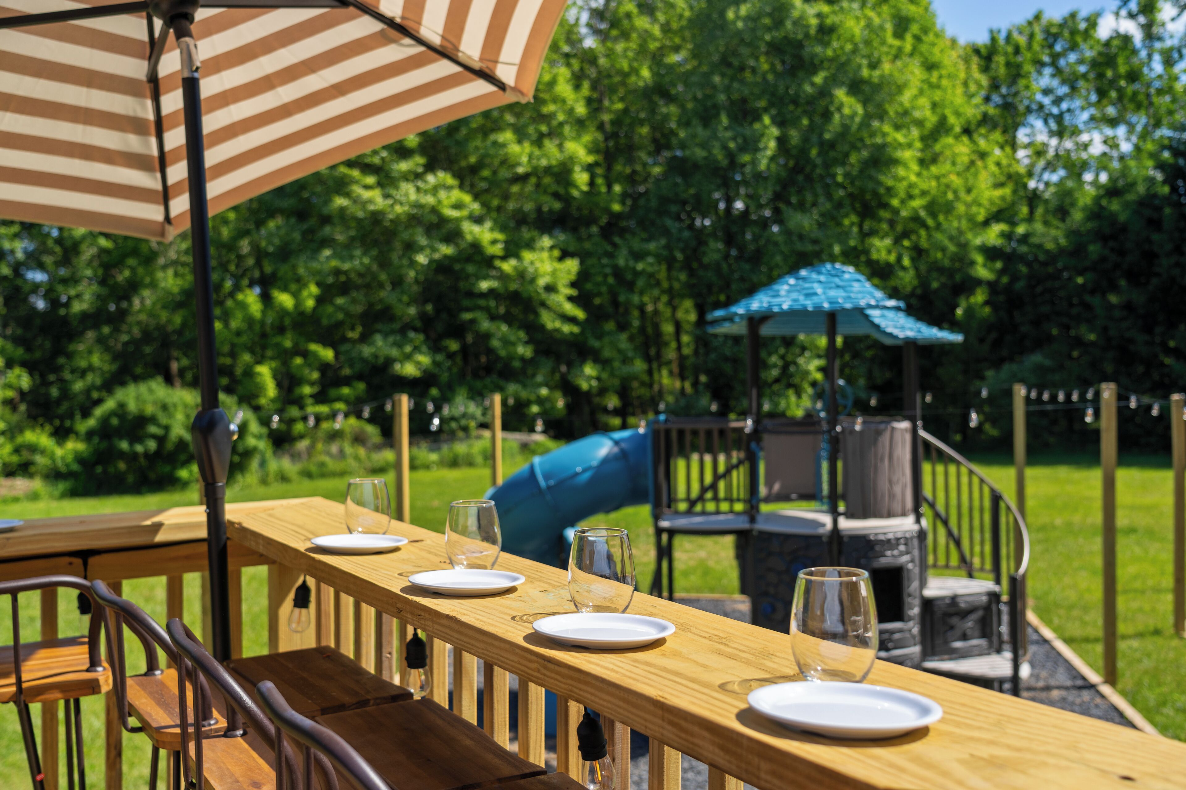 Un espace extérieur aménagé pour les repas, avec des tables et des chaises en bois, un parasol à rayures et, au fond, une aire de jeux.