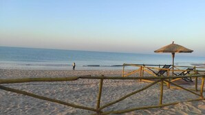 Una playa con una cerca de bambú, una sombrilla de paja y sillas de descanso.