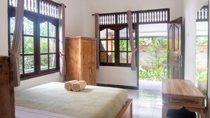 A bedroom with a bed, wooden wardrobe, and a view of greenery through open windows. - Sandora Homestay by Supala (Ubud)