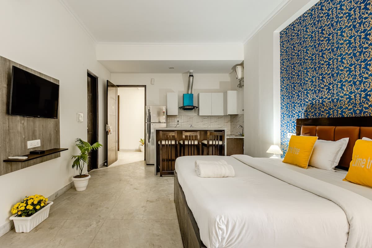 A modern hotel room with a large bed, a TV mounted on the wall, a kitchen area with cabinets, and a patterned wall.