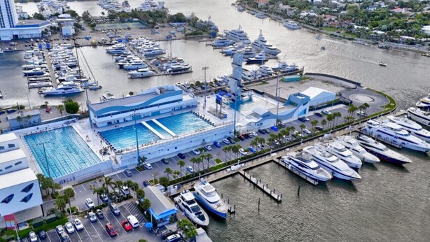 Eine Vogelperspektive einer Marina mit zahlreichen vertäuten Yachten, einem großen Schwimmbecken und einem Gebäude mit blauem Dach.