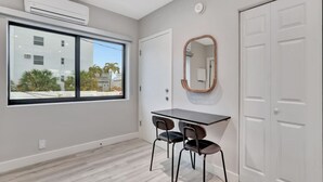 Ein Zimmer mit einem großen Fenster, einem Schreibtisch mit Stühlen, einem Spiegel und einem Kleiderschrank.