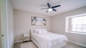 A bedroom with a bed, a nightstand, a ceiling fan, a window, and a painting on the wall.