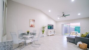 A modern living room with a gray sofa, a dining area with a glass table and chairs, and a TV on a white cabinet.