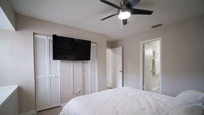 Ein Schlafzimmer mit einem Bett, einem Deckenventilator, einem an der Wand befestigten Fernseher und einer Tür zu einem anderen Raum.