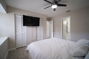 Ein Schlafzimmer mit einem Bett, einem Deckenventilator, einem an der Wand befestigten Fernseher und einer Tür zu einem anderen Raum.