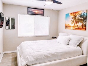 Ein Schlafzimmer mit einem Bett, einem Deckenventilator, einem Fenster mit Jalousien, einem kleinen Tisch und einem Bild an der Wand.